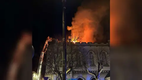 Aimee Gardner Flames and smoke seen against the night sky and firefighters alongside the burning building on an aerial platform, spraying water on the flames.