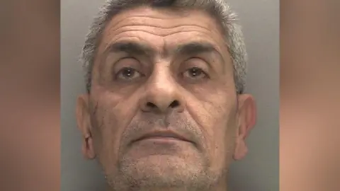 West Midlands Police Dhiya Al Maamoury, who stares at the camera, with grey hair and light stubble. The picture is a close-up of his face, which is in front of a grey background.