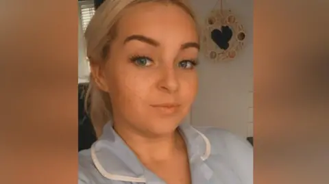 Jess Thorpe Jessica Thorpe in her nurse uniform, slightly smiling at the camera, with a snapchat filter layered over the top, giving her freckles.