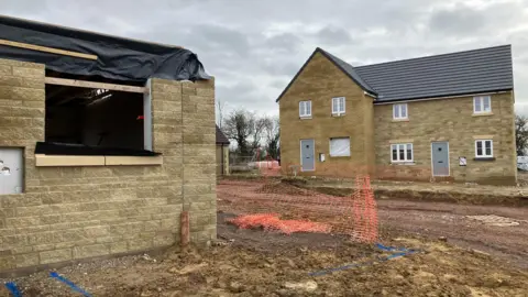 A building site with a newly-built pair of yellow stone semi-detached houses in the background with a half-built house in the foreground with no roof or windows