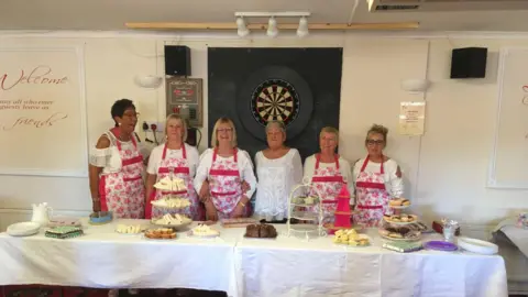 Alan Baldwin A group of women are behind a long table covered with cakes, sandwiches and afternoon tea stands inside a community hall, with a dartboard on the wall behind them. Several people wear matching patterned aprons, and trays of food and plates are laid out across the table.