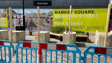 Suki Somal/BBC Barriers in front of a building with large concrete block and wooden poles behind them.