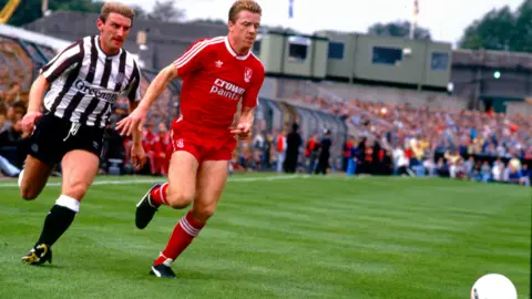 Steve Nicol with all-red football kit is chasing a ball pursued by a Newcastle player in black and white stripes with black shorts and black socks with white tops.