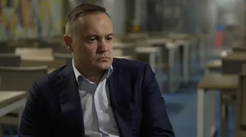 Matthew Goddard/BBC A man sits in a blue jacket in a darkened classroom