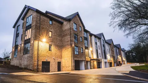 A large brown-brick apartment complex, with outside lights on in the middle of the front of the building. There are pitched roofs along the complex with some trees to the right.