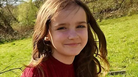 Aria Thorpe wearing a burgundy jumper, sitting outside on a grassy hill surrounded by trees on a sunny day. She has long wavy brown hair and is smiling at the camera.