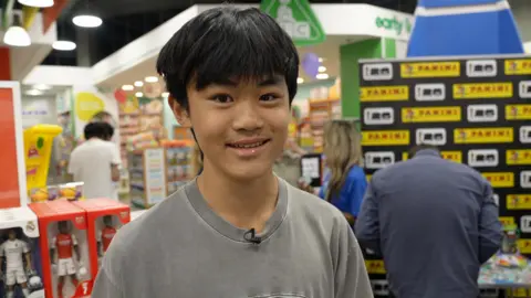 A teenage boy with short black hair, with a fringe, smiling at the camera wearing a grey T-shirt and is standing in front of a Panini sign.