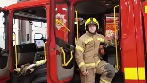 Barry Martin stands at a fire engine, dressed in his firefighting uniform with yellow helmet on