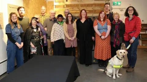 CamSight A group of people, standing looking at the camera, at a listening party at Babylon Arts. It shows a woman with a guide dog, a woman, dressed in a costume with a stick, and a mixture of men and women. 