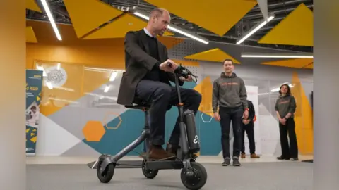 PA Prince William on a stream-lined mobility scooter in a large conference hall, with several people in a uniform of grey hoodie and trousers milling in the background. 