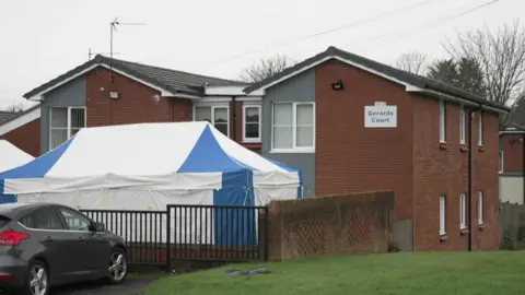 A two storey block of flats with a forensic tent over the garden area outside. A sign says Gerards Court.