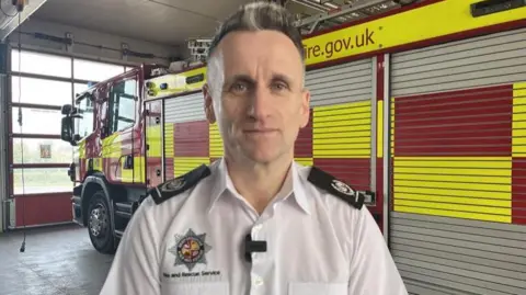 Nicola Hasler/BBC Wayne Swales is in a white shirt uniform standing in front of a fire engine. He is inside a station, with a large garage-type door in the background.