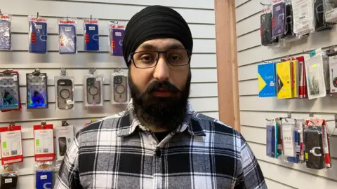 A man with a black Dastaar on his head. He wears black glasses, a black t-shirt and a black and white checked shirt. Behind him on the wall are rows of phone cases and accessories
