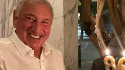 David Berman with white hair wearing a white shirt is smiling. He is in a restaurant and a cake with candles with the number 86 burning. 