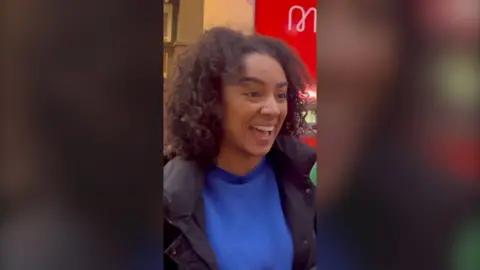 A woman with curly black hair laughs in the street while talking into a mic