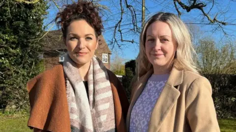 Amira Stallion and Amber Bradly stand side by side with a hedge behind them. Amira is wearing an orange coat and patterned scarf. Her brown curly hair is tied in a ponytail. Amber is wearing a cream coat and has blonde mid length hair. 