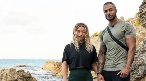 StudioLambert/BBC Tyler West and Molly Rainford hold hands and smile stood on rocks by the shore of the sea.
