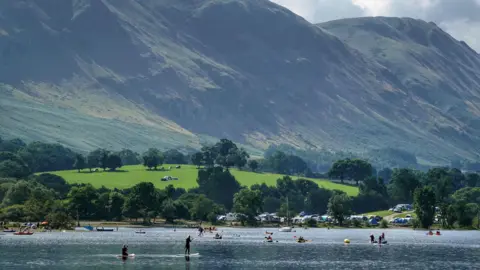 Ullswater in the Lake District, with the high fells on the horizon. There are several people paddleboarding and swimming in the lake. It is a summer's day, where there is bright green grass and several trees. 