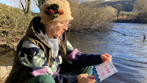 Kathy Oakwood sitting near river Tawe, holding a test tube and looking at a testing water quality pamphlet. 