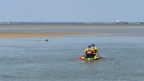 Wirral Coastguard Rescue Team Two men dressed in yellow and black waterproof clothing and blue helmets are rowing a small inflatable yellow craft towards a dog that it is in the water near to a sandbank. A large ship can be seen in the distance.