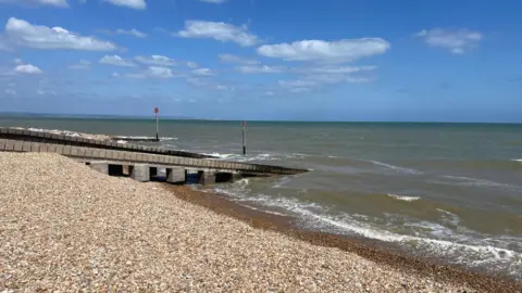 A pebble beach. The seawater is dark and muddy. The sky is bright blue. 