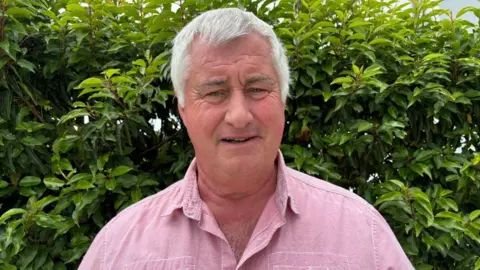 A man with white hair, wearing a pink shirt looking at the camera