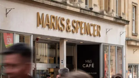 Marks & Spencer sign above a shop in gold lettering. People are walking past the shop and are blurry.