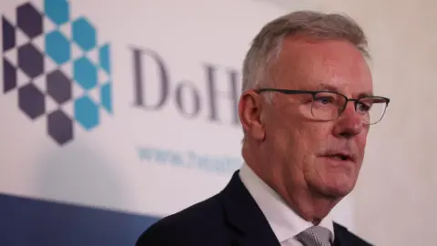PA Media Mike Nesbitt. He has short grey hair, wearing glasses, a black blazer, white shirt and grey tie. He is standing in front of Department of Health signage.