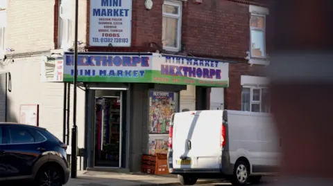 The exterior of a small neighbourhood convenience store located on a street lined with brick buildings. The shop is called “Hexthorpe Mini Market,” as displayed prominently on large signage above the entrance. The sign features bold, colourful text- mainly blue, green, and red - and includes additional details such as “Grocery, Meat, Bakery, Fruit & Veg, Dairy.”