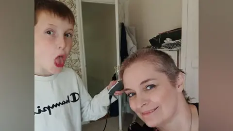 Louise Palmer A boy with short brown hair is sticking his tongue out at the camera while holding some hair clippers at his mum's head. Louise has a partially shaved head and is smiling at the camera. They are in a carpeted room with patterned wallpaper.