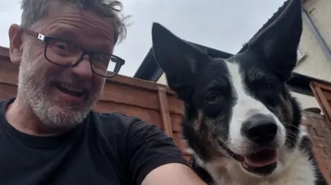 Chris Durham, a man with a grey beard, glasses and a dog are stood outside next to a brown wooden garden fence