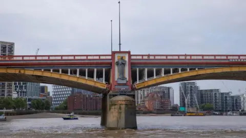 A red and yellow bridge with a statue positioned on one of the supports. The background shows a skyline of high rise buildings. 