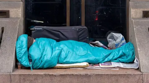 Getty Images Homeless person in a blue sleeping bag on folded out newspapers on a doorstep. There is a black suitcase and supermarket plastic bag in the background