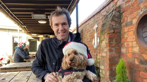 Worcester Parkrun A man smiles at the camera while holding a blond dog in a Santa hat.
