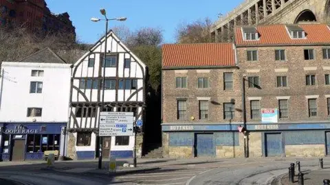 Andrew Curtis/Geograpgh The Cooperage in 2009. It is a white four-storey house with timber frame next to a flight of stairs. The ground floor's facade on the left side of the building is blue and has signage reading The Cooperage. A row of brick buildings is immediately next to the flight of stairs. Newcastle's High Level bridge is rising above in the distance.