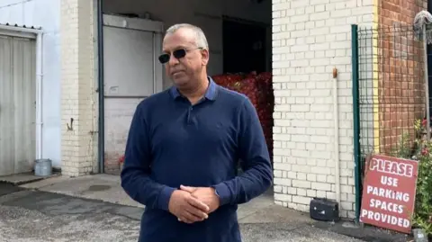 VICTORIA SCHEER/BBC Mojid Kha, owner of Sunrise Catering Supplies, stood in front of his business, wearing sunglasses and a dark blue top, with his hands together and a serious look on his face