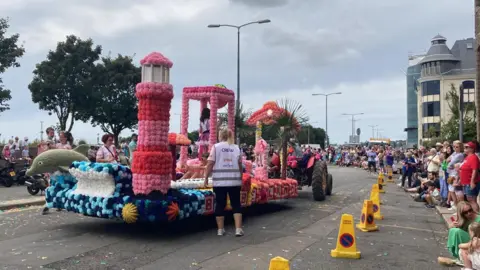 A carnival float covered in pink and blue flowers on a road. The float has a decorative dolphin, seashells and a lighthouse. Lots of people are sitting and standing on the pavement to watch. There are yellow traffic cones along the edge of the road. It's a cloudy day.
