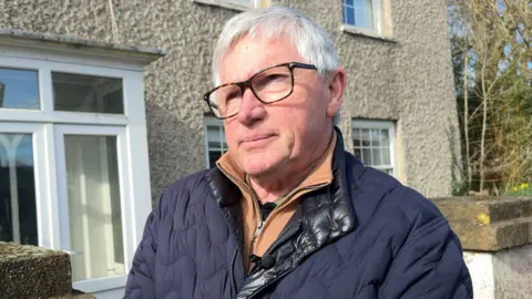 Senior Constable of St Martin Dave Beausire stands in front of his parish's rectory. He has square-rimmed glasses on and is wearing a navy blue coat and sandy coloured jumper.