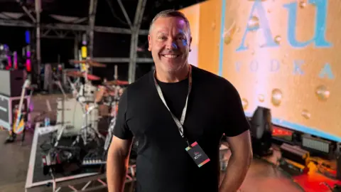 A man in a black t shirt with a stage pass lanyard standing on the stage. The drums are in the background and a guitar.