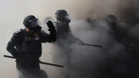 Officers wear riot gear and hold batons as they walk through smoke.