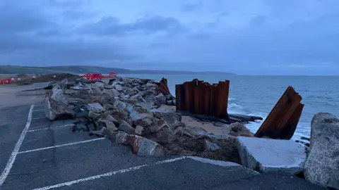 Boulders placed on the sand next to the collapsed road. The sea is lapping on the sand next to the damaged road.
