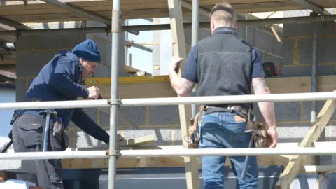 Two men are standing on scaffolding looking at a wall made of block. The older man is sawing a piece of wood while the younger one is watching and holding a long piece of timber. They are both wearing tool belts.