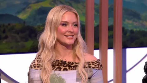 Zara Larsson sits in The One Show studio - she has long blonde hair and wears a top with leopard print edge