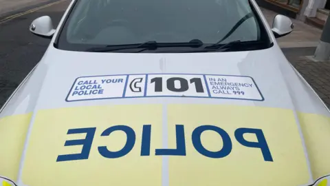 The bonnet of a yellow and white police car with the word police written backwards in large blue letters and a label telling people to call 101