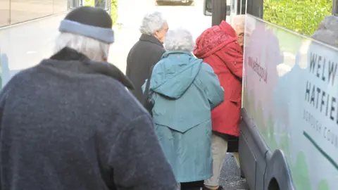 The backs of four elderly people who are queueing to get on board a bus.