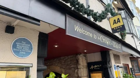 This photo includes the exterior of the Crown at Wells pub. The walls are creamed coloured with bay windows. Their is a blue plaque on the building which reads: The International Blockbuster Hot Fuzz filmed at The Crown 2006