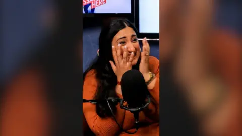 A woman wearing an orange jumper smiles and holds her hands up to her face.