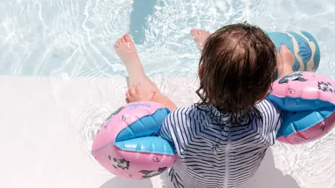 A toddler with dark, wet hair, who is wearing a black and white striped swimsuit and pink and blue armbands, sits on the edge of a swimming pool with their feet in the water