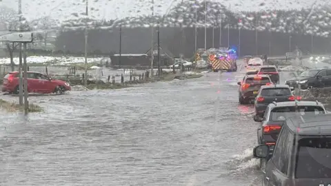 Traffic queues along a flooded road as cars drive through standing water, with emergency vehicles flashing blue lights in the distance during heavy rain.