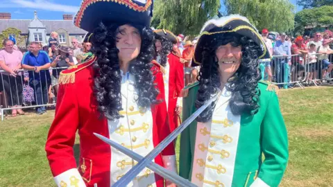Two men are dressed up in Jacobean era costumes. Both are wearing long curly wigs. One is in red and the other is in green. Both are holding their swords up. A crowd is behind them behind a fence. 
